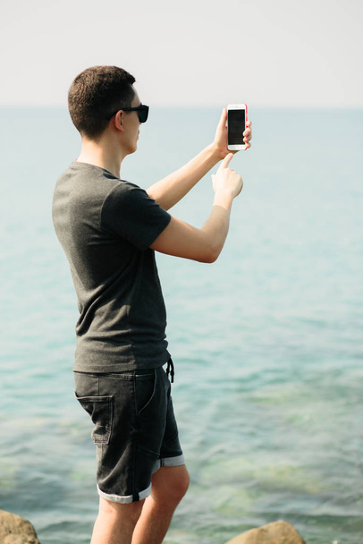 ein junger Mann in T-Shirt und kurzen Hosen auf großen Felsen, der mit seinem Smartphone einen malerischen Ort an der Küste fotografiert - Foto, Bild