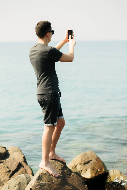a young guy in a t-shirt and shorts on large rocks, taking pictures of a picturesque place on the sea coast on a smartphone - Photo, Image