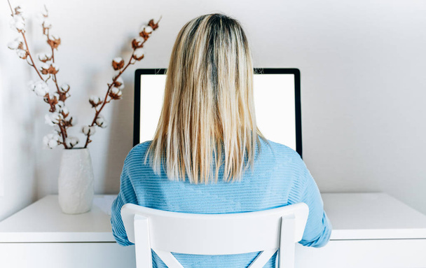 Rear view of businesswoman using desktop computer in home office. Back view of young woman in blue sweater working indoor at modern office on computer with blank screen for your text message, content - Foto, Imagen