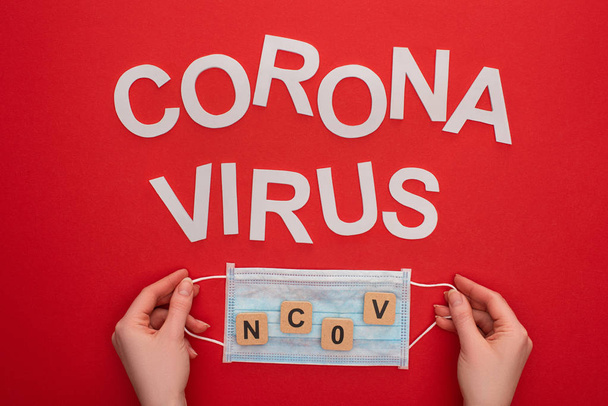 Bovenaanzicht van vrouw met medisch masker met ncov tekens op houten blokjes en coronavirus letters op rode achtergrond - Foto, afbeelding