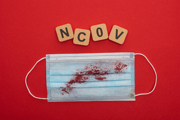 Draufsicht auf medizinische Maske mit Blut und ncov Buchstaben auf Holzklötzen auf rotem Hintergrund  - Foto, Bild
