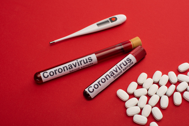  Pillen, thermometers en reageerbuizen met bloedmonsters en coronavirusbelettering op rood oppervlak - Foto, afbeelding