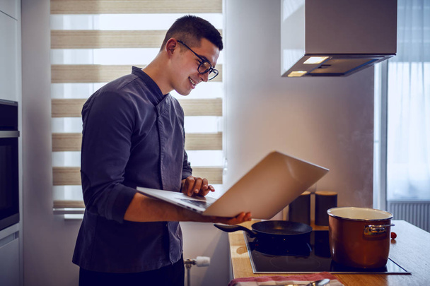 Chef caucasiano sorridente em uniforme e com óculos usando olhar para a receita no laptop enquanto está ao lado do fogão. No fogão são panela de cozinha e panela
. - Foto, Imagem