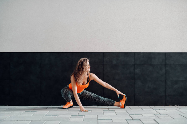 Beautiful slim caucasian sporty woman in sportswear and with curly hair stretching her leg before running. In background is black and white wall. - Valokuva, kuva