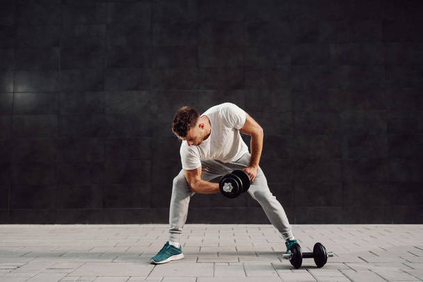 Full length of attractive muscular caucasian blond bearded man in tracksuit and t-shirt lifting dumbbells while standing in front of gray wall. - Foto, immagini