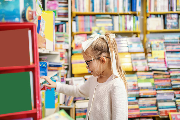 Vista lateral da encantadora menina loira caucasiana com rabo de cavalo e com óculos em pé na livraria e escolhendo o que ler em seguida
. - Foto, Imagem