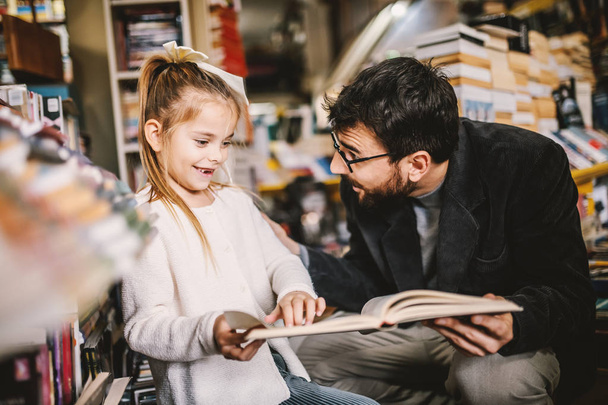 Adorable little caucasian blond girl without two teeth reading book with his caring father in bookstore. - Photo, Image