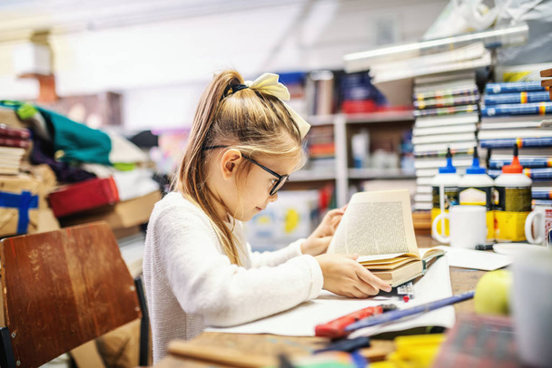 Adorable caucasian blond girl with ponytail and with eyeglasses sitting in bookstore and reading exciting book. - Photo, Image