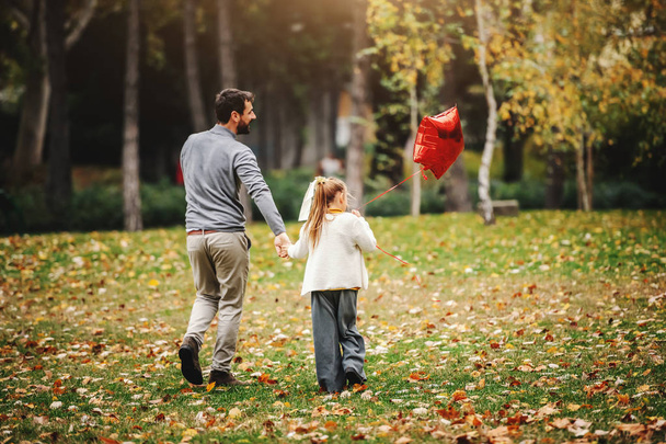 entzückendes kleines blondes Mädchen, das Luftballons hält und mit ihrem Vater auf der Wiese spazieren geht. Herbstzeit. Qualität Familienzeit. - Foto, Bild