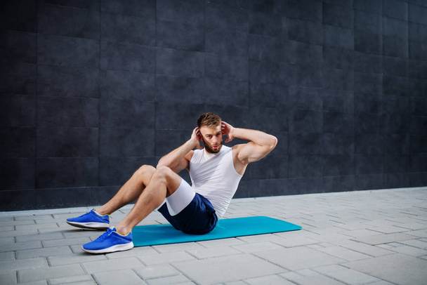 Young muscular caucasian handsome man in shorts and t-shirt doing abs on mat outdoors. In background is gray wall. - 写真・画像