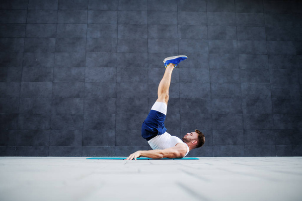 Vista lateral do homem caucasiano muscular forte em shorts e camiseta fazendo exercício castiçal no tapete ao ar livre. No fundo é parede cinza
. - Foto, Imagem