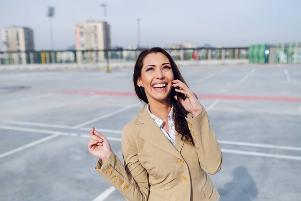 Prachtige Kaukasische brunette gekleed in beige jas staan op de parkeerplaats en met behulp van smart phone voor het bellen van taxi. - Foto, afbeelding
