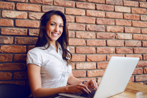 Happy pretty caucasian brunette sitting in cafe and using laptop. Hands are on keyboard. In background is brick wall. - Fotó, kép