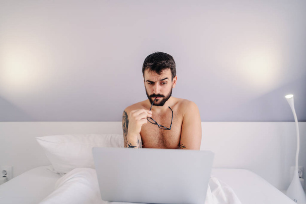 młody biały koszule brodaty mężczyzna siedzi na łóżku w sypialni i za pomocą laptopa. - Zdjęcie, obraz