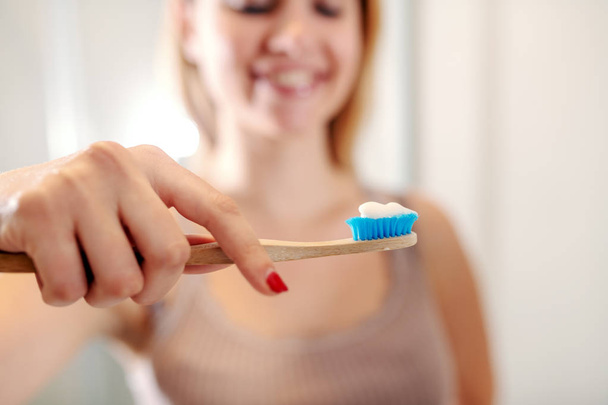 Beautiful smiling caucasian woman standing in bathroom and holding toothbrush with toothpaste. Morning routine. Selective focus on toothbrush. - Photo, Image