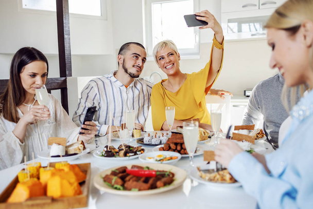 Grupa wielokulturowych przyjaciół je zdrowy lunch w domu. Dwóch z nich robi sobie selfie.. - Zdjęcie, obraz