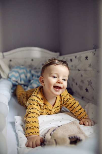 Cute cheerful smiling blond toddler with beautiful blue eyes lying on stomach in his crib in the morning and waking up. - Foto, Bild