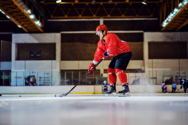 Longitud completa de intrépido jugador de hockey patinando y preparándose para golpear disco. Interior del salón. Deportes de invierno
. - Foto, Imagen