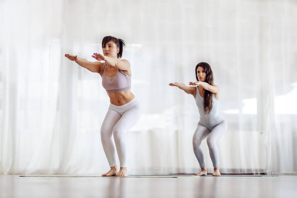 Twee mooie jonge vrouwen met bruin haar en gezonde gewoonten staan op de mat op blote voeten in ongemakkelijke yoga positie. Yoga studio interieur. Totale lengte. - Foto, afbeelding
