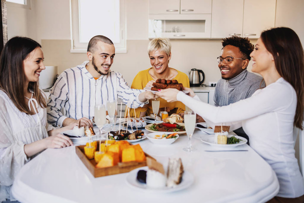 Group of multicultural happy friends sitting at dinning table and having lunch at home. Woman holding plate with meat rolls and passing to a guy. - Zdjęcie, obraz