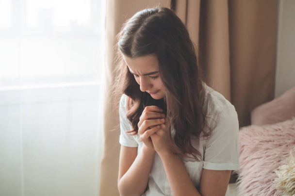 Genç kız gözlerini kapadı, oturma odasında dua ediyordu. İnanç için dua kavramına sarılmış eller. - Fotoğraf, Görsel