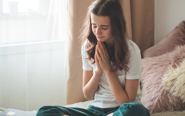 Η έφηβη έκλεισε τα μάτια της, προσευχόμενη στο σαλόνι. Χέρια διπλωμένα στην ιδέα της προσευχής για πίστη - Φωτογραφία, εικόνα