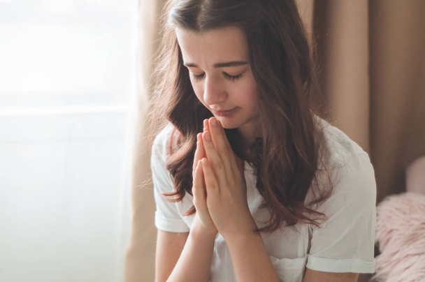 Η έφηβη έκλεισε τα μάτια της, προσευχόμενη στο σαλόνι. Χέρια διπλωμένα στην ιδέα της προσευχής για πίστη - Φωτογραφία, εικόνα