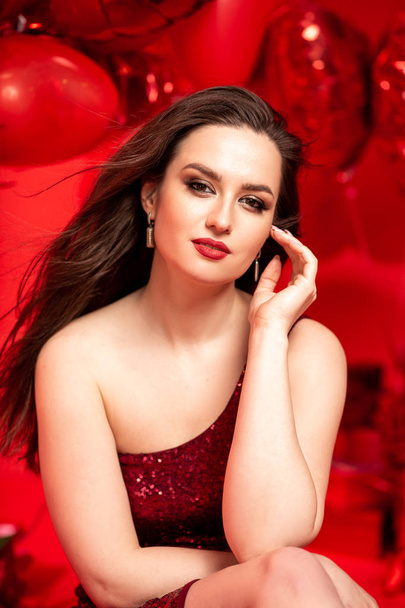 Beautiful young woman in red evening dress posing over red background with big heart shape balloons. Happy Valentine's Day concept - Fotoğraf, Görsel