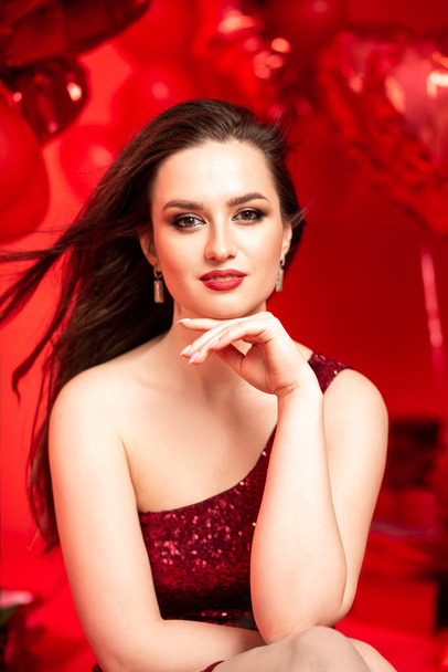 Beautiful young woman in red evening dress posing over red background with big heart shape balloons. Happy Valentine's Day concept - Фото, зображення