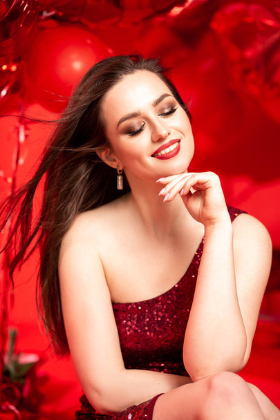 Beautiful young woman in red evening dress posing over red background with big heart shape balloons. Happy Valentine's Day concept - Fotografie, Obrázek