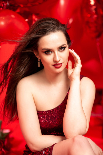 Beautiful young woman in red evening dress posing over red background with big heart shape balloons. Happy Valentine's Day concept - Fotó, kép