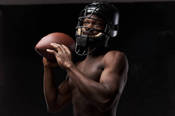 portrait photo of dark-skinned young man with nude torso on a dark background he has a rugby helmet on head and holds a rugby ball up in his arm - Фото, зображення