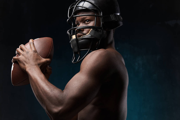 portrait photo of dark-skinned young man with nude torso on a dark background he has a rugby helmet on head and wants to throw a rugby ball  - Fotoğraf, Görsel