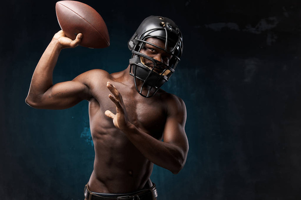portrait photo of dark-skinned young man with nude torso on a dark background he has a rugby helmet on head and holds a rugby ball up in his arm - Foto, Imagem