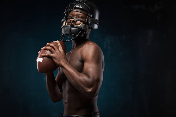 portrait photo of dark-skinned young man with nude torso on a dark background he has a rugby helmet on head, holds a rugby ball in his arms - Zdjęcie, obraz