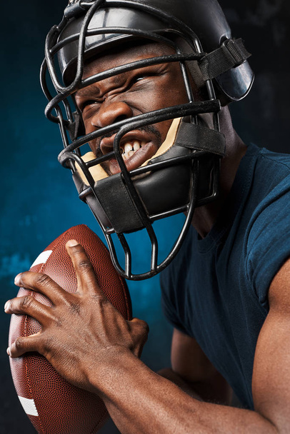 portrait photo of dark-skinned young man on a dark background wearing a t-shirt, rugby helmet and holds rugby ball in his arms he has anger on his face - Foto, Imagem
