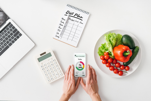 Top view of woman holding smartphone near calculator, notebook and vegetables on white background - 写真・画像