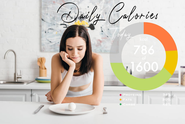 Pensive sportswoman looking at egg on kitchen table near daily calories illustration - Photo, Image