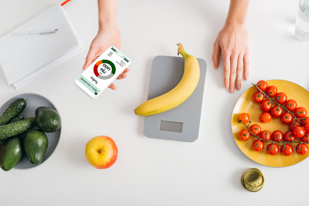 Top view of girl holding smartphone with calorie counting app while weighing banana on kitchen table near fresh vegetables - Photo, Image