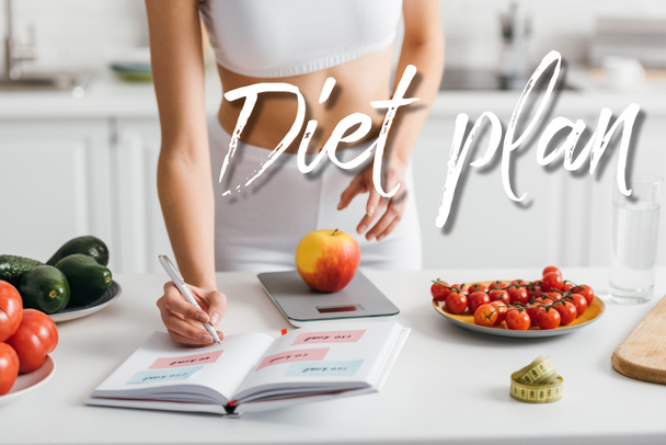 Cropped view of fit sportswoman writing calories while weighing apple on kitchen table, diet plan illustration - Photo, Image
