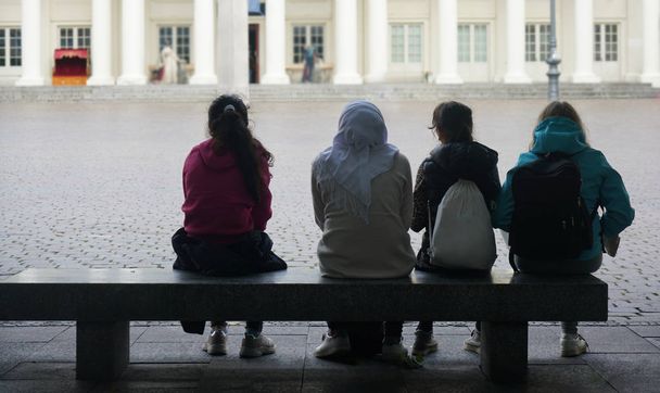 back, rear view of school girls sitting on stone bench against school building. asian and caucasian  teenagers waiting together - Foto, Bild
