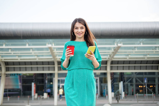 Jeune femme heureuse tient un smartphone et sourit. Café chaud dans un verre en plastique. Robe verte. Le concept d'entreprise, de style de vie et de technologie
. - Photo, image