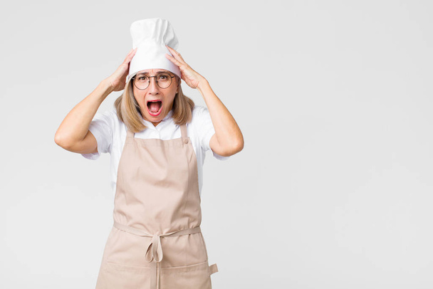 middle age baker woman looking excited and surprised, open-mouthed with both hands on head, feeling like a lucky winner against flat wall - Foto, Imagen