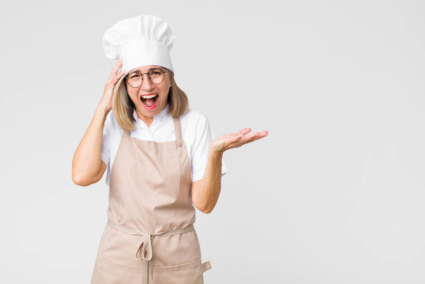 middle age baker woman laughing, looking happy, positive and surprised, realizing a great idea pointing to lateral copy space against flat wall - Φωτογραφία, εικόνα