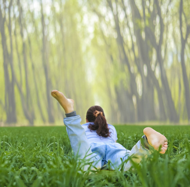 barefoot young woman lying in fresh green grass and reading a book. back view.woman lying on spring green grass. summer time season. unrecognizable person. springtime in park. - Photo, Image