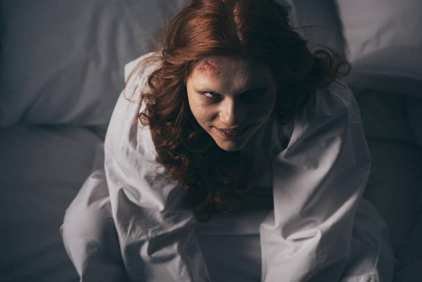 bovenaanzicht van demonische lachende vrouw in nachtjapon zittend in bed - Foto, afbeelding
