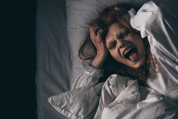 Вид сверху одержимой девушки в ночном халате, кричащей, лежа в постели
 - Фото, изображение