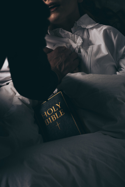 καλλιεργημένη άποψη του εξορκιστή με Αγία Γραφή κρατώντας τα χέρια με δαιμονική εμμονή κορίτσι στο κρεβάτι  - Φωτογραφία, εικόνα