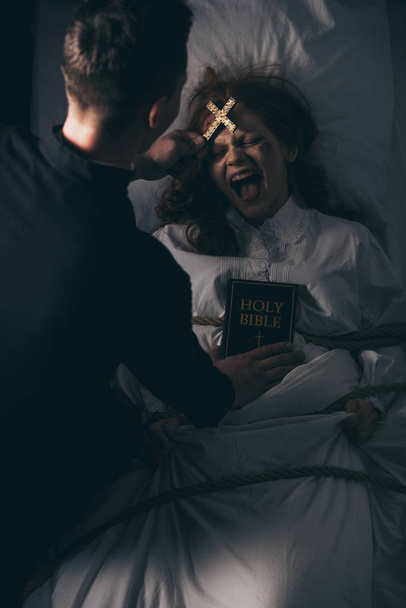 egzorcystka z biblią i krzyżem stojąca nad opętaną wrzeszczącą dziewczyną w łóżku - Zdjęcie, obraz