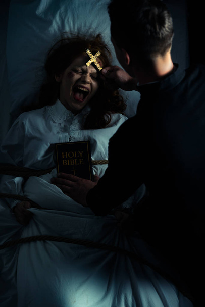 male exorcist with bible and cross standing over demonic screaming girl in bed - Photo, Image